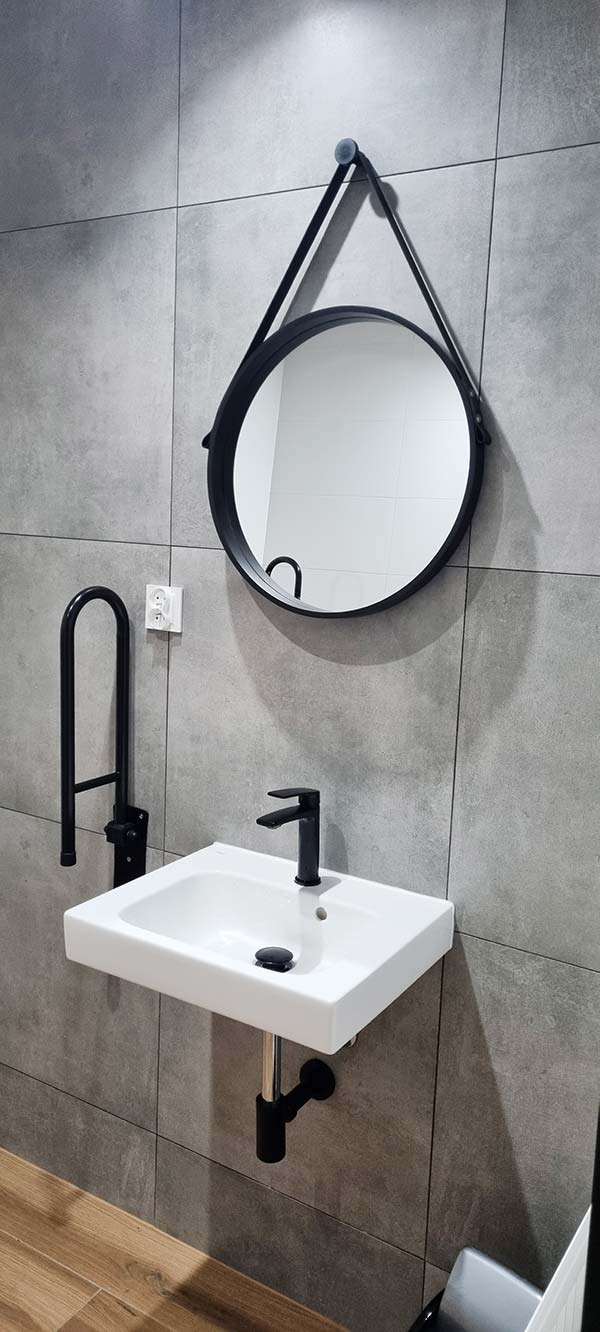 Łazienka w gabinecie, usprawnienia dla niepełnosprawnych, biały zlew oraz okrągłe lustro w czarnej metalowej ramie.